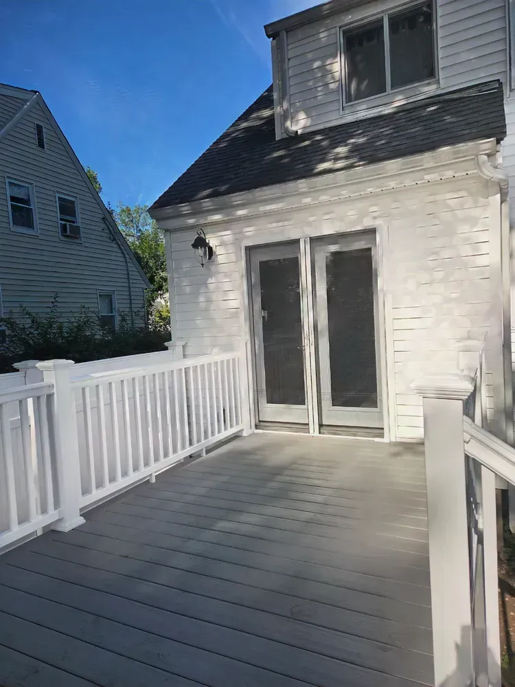 White deck with railing leading to a sliding glass door on a two-story house under a blue sky