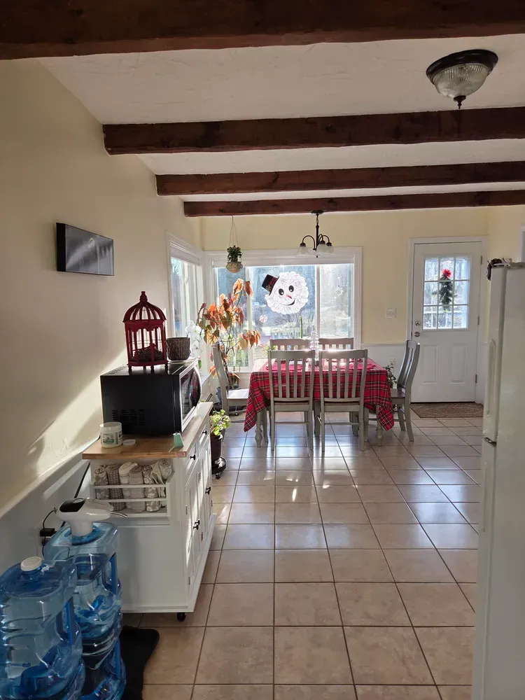 Small dining room with wooden ceiling beams, tiled floor, table set by a bright window, and water bottles by a cabinet