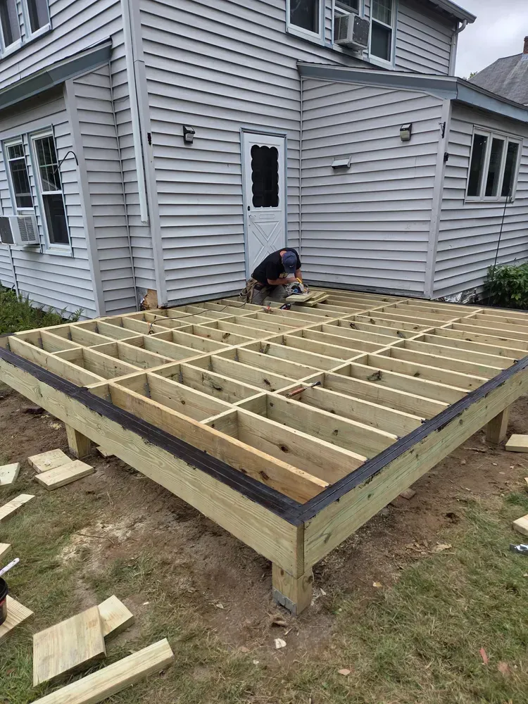 Worker framing a wooden deck beside a gray house under construction.