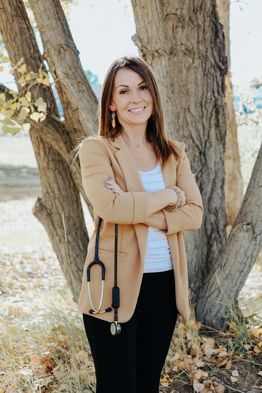 Woman in tan blazer smiles, stands outdoors with arms crossed, stethoscope around neck.