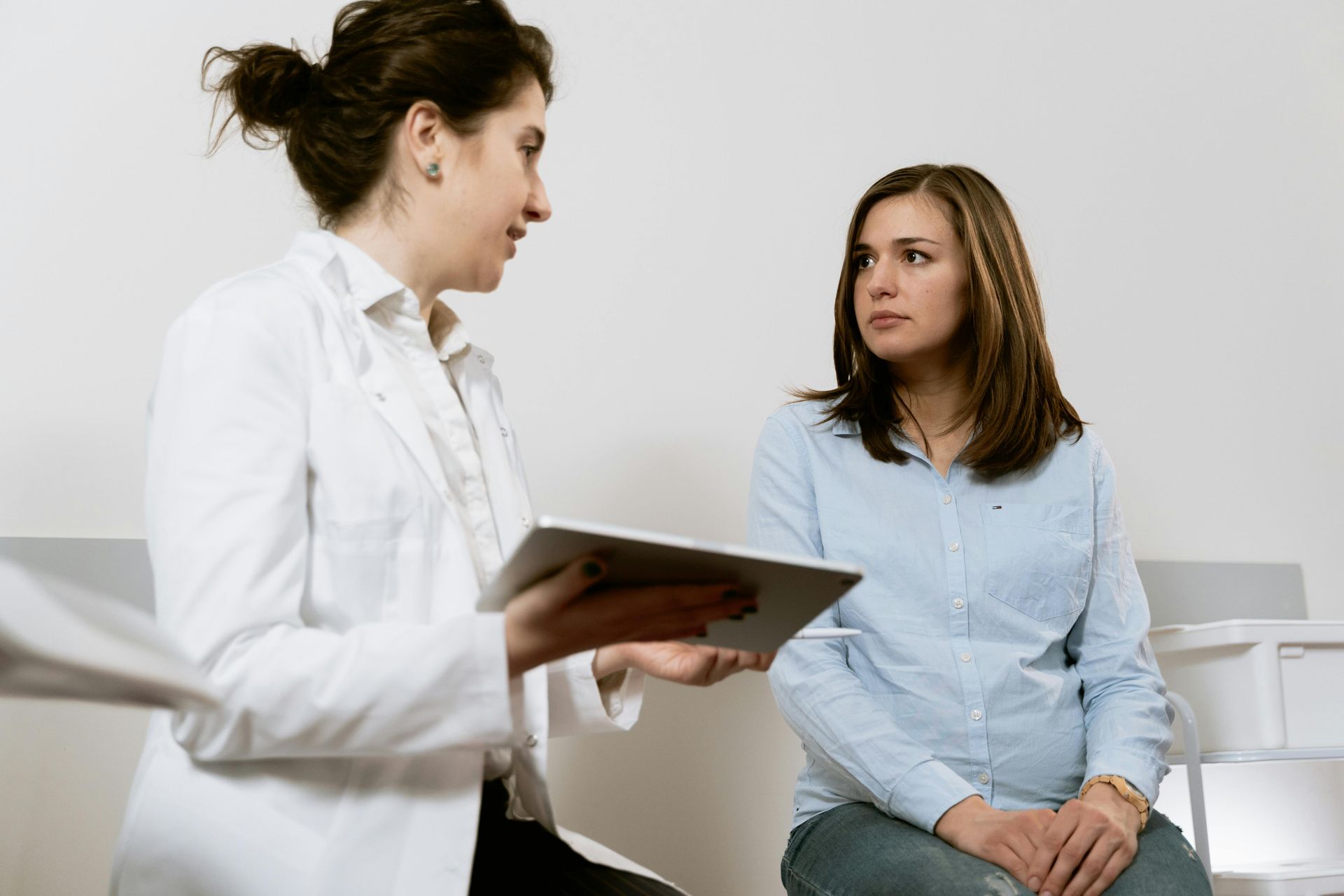 Doctor in white coat reviews tablet with seated patient in light blue shirt.