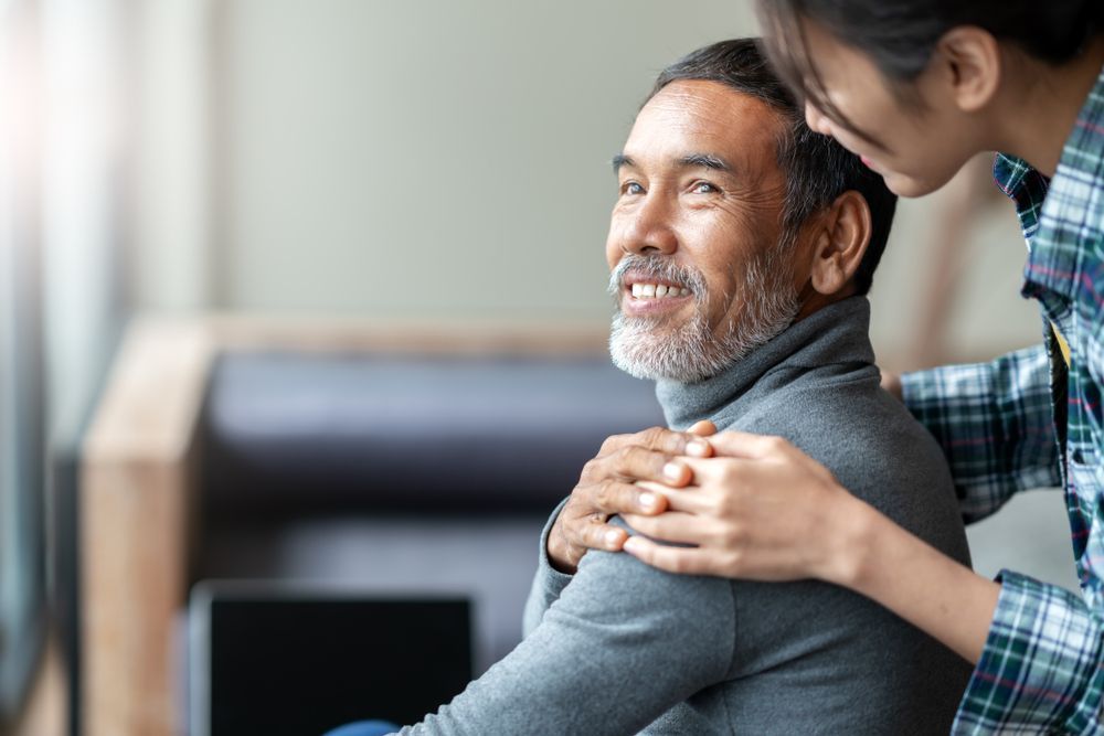 Man smiles while a person's hands rest on his shoulders. Indoors, natural light.
