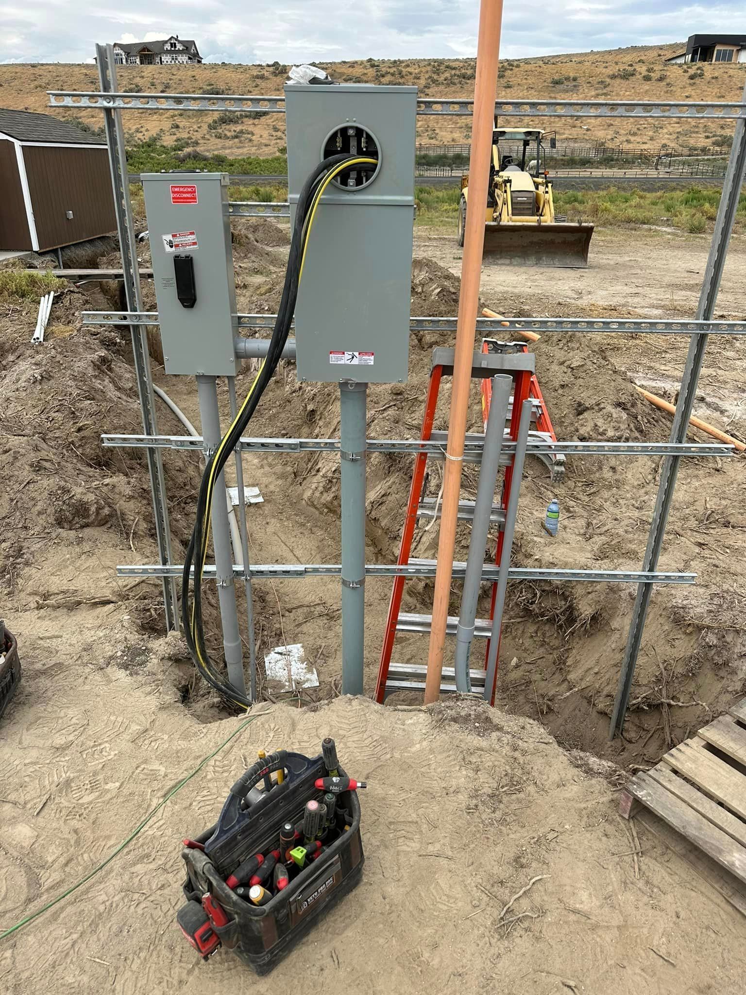 Electrical work in progress at a construction site: gray metal boxes, yellow/black cable, ladder, tools, soil, and a backhoe.