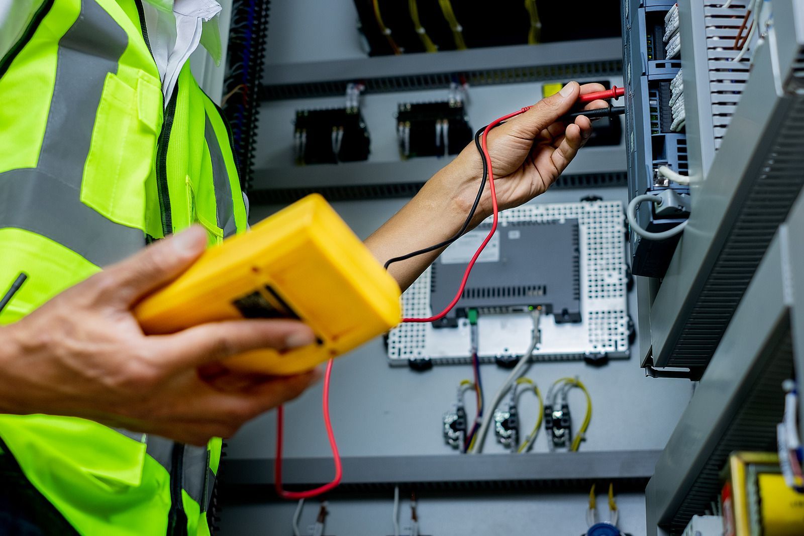 Electrician using a multimeter to test electrical equipment inside a panel; wearing a safety vest.