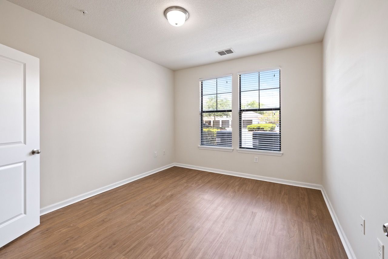 Empty beige bedroom with two windows, blinds, and wood-look flooring.