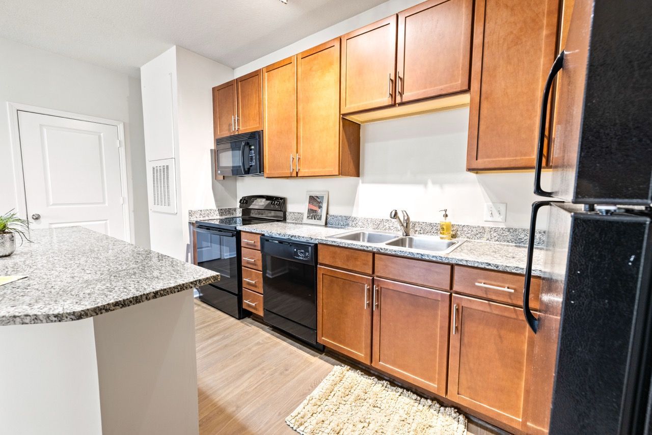 Modern apartment kitchen with granite countertops, wooden cabinets, black appliances, and a double sink.