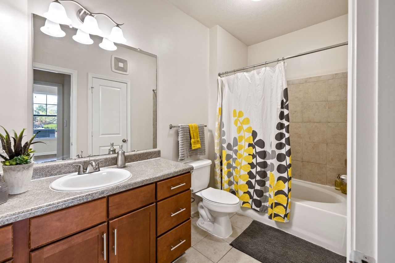Apartment bathroom with a single vanity, mirror, toilet, and tub/shower with a yellow-gray patterned curtain.