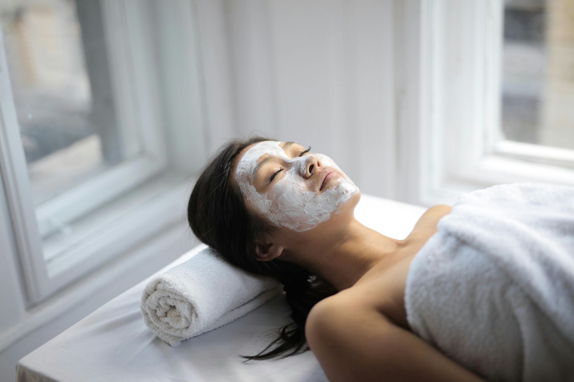 Woman relaxing with a facial mask at spa, eyes closed, white towel, near window.
