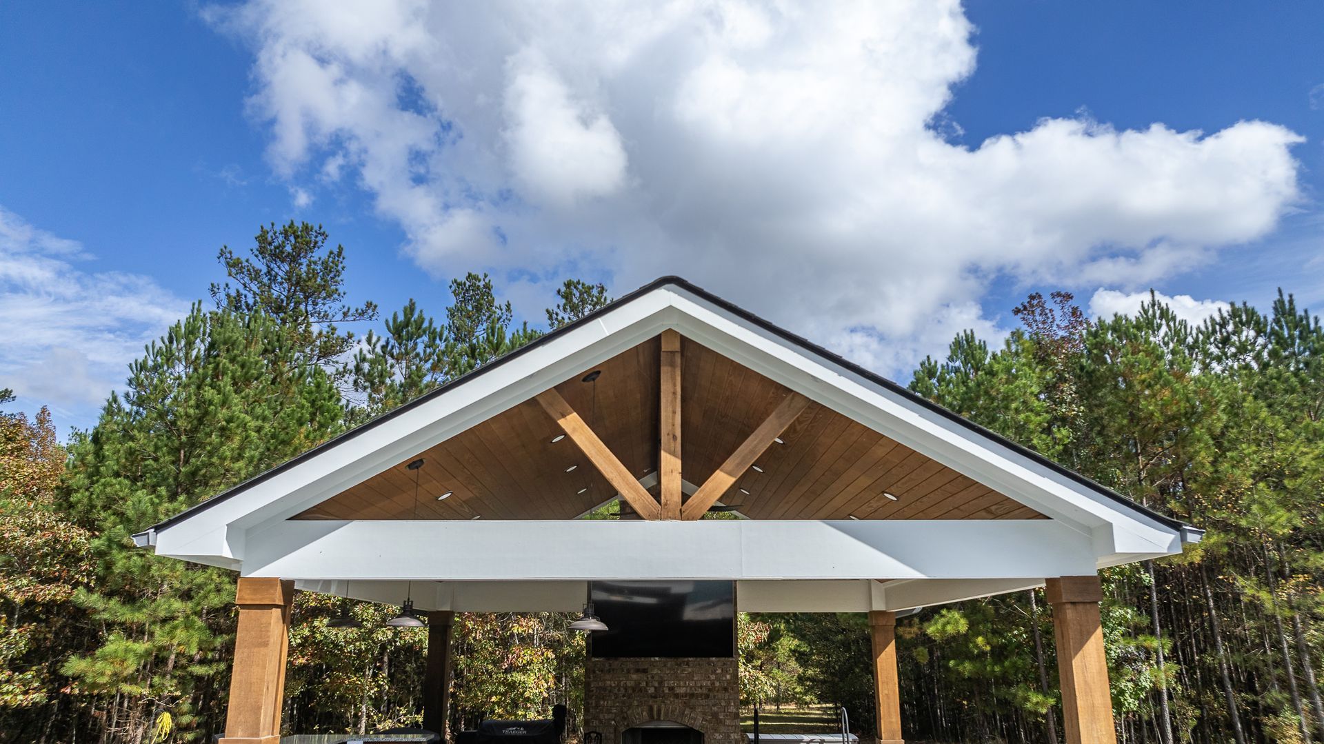 A pavilion with a wooden roof and a fireplace in the middle of a forest.