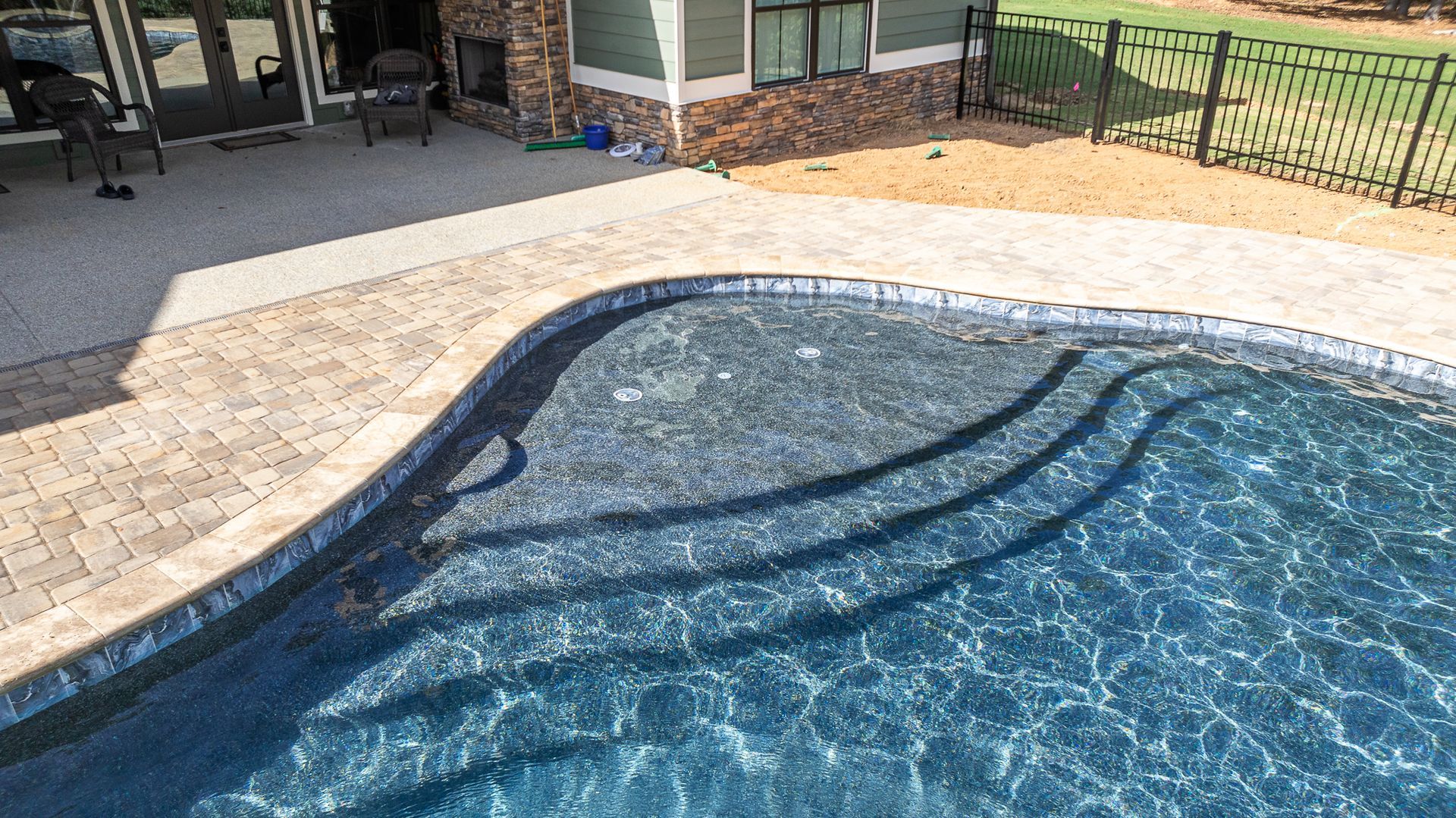 A large swimming pool is surrounded by a brick patio and a fence.