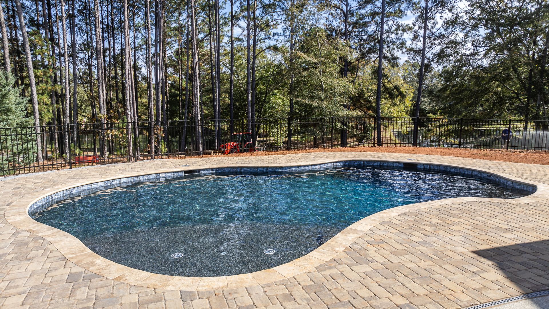 A large swimming pool is surrounded by trees and a brick patio.