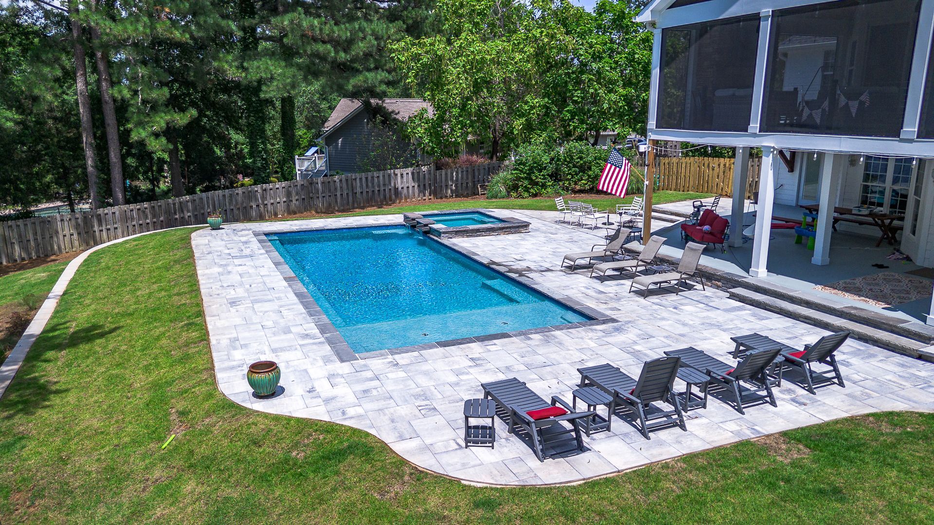 Backyard with pool, hot tub, patio furniture, and a house. Blue water, green grass, and blue sky.