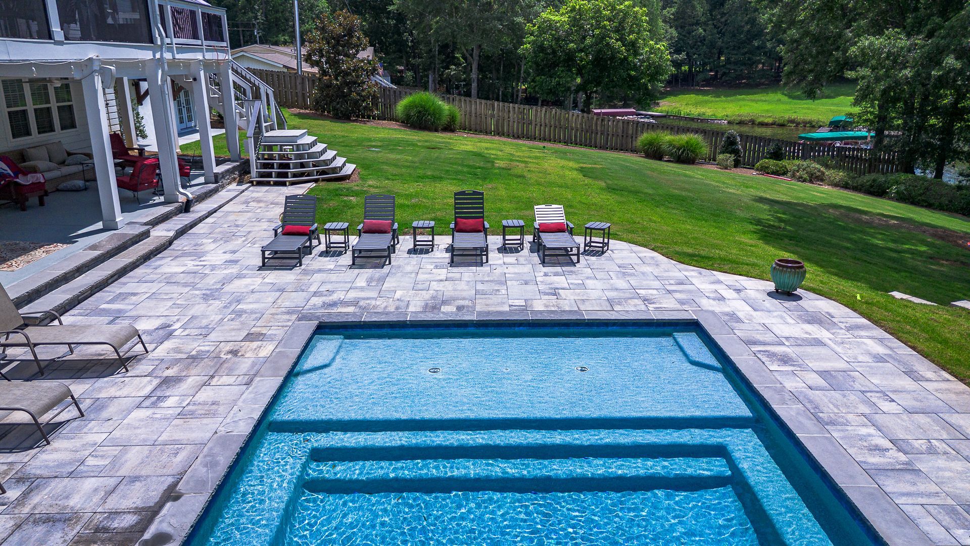 Backyard with pool, lounge chairs on patio, green grass, and a white house.
