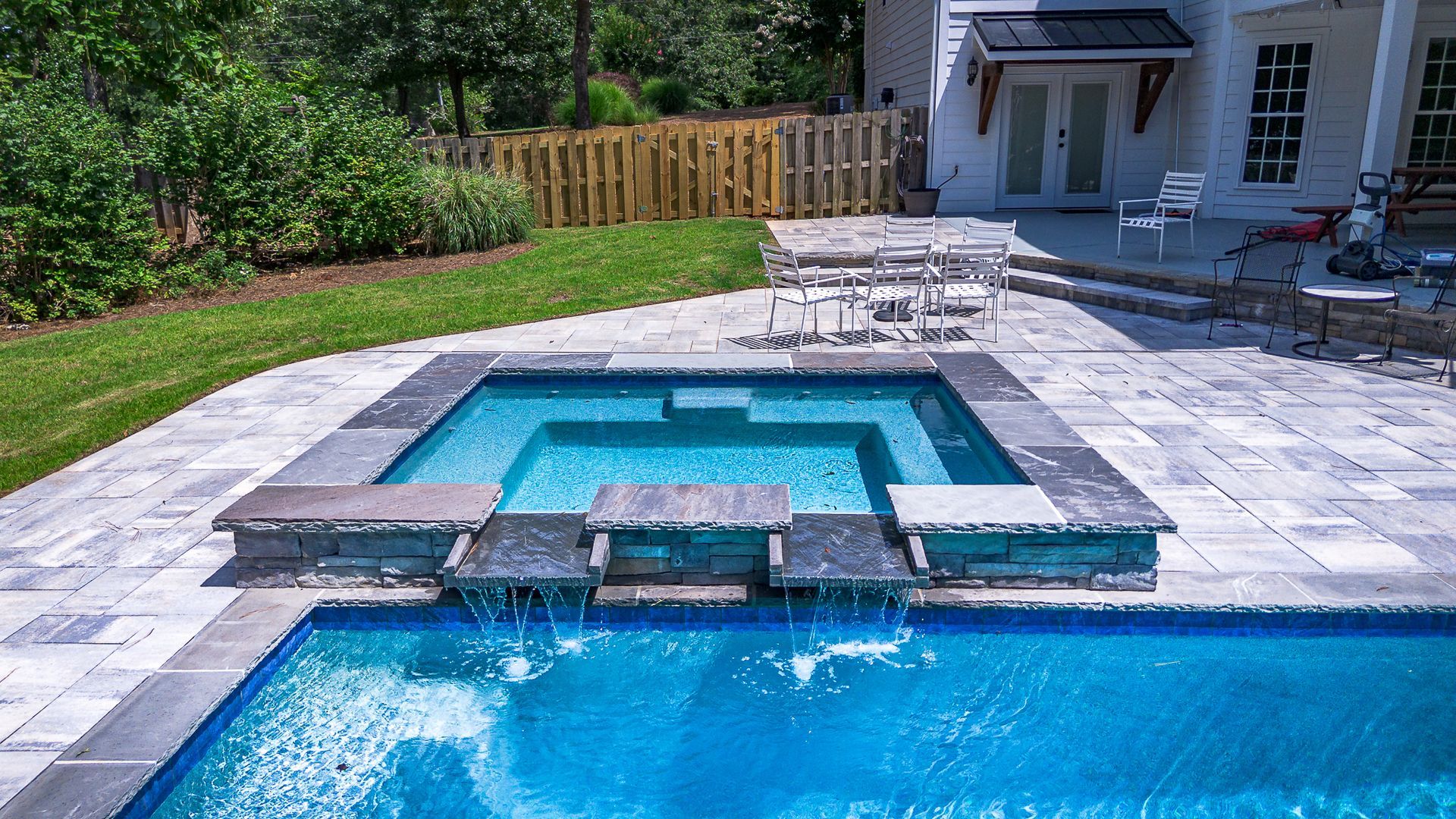 A rectangular pool with a raised spa, waterfall, and patio, next to a house and yard.