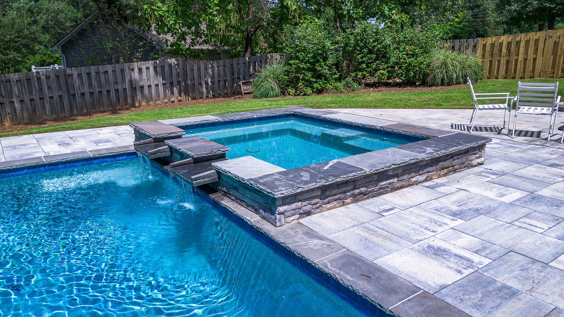Swimming pool with a hot tub, both surrounded by gray pavers. Blue water, trees in the background.