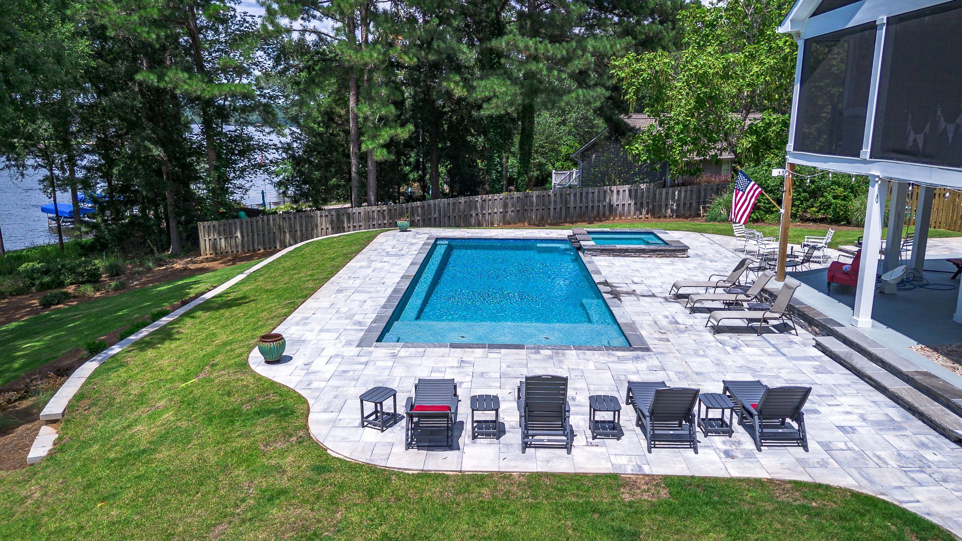 Backyard with rectangular pool, lounge chairs, hot tub, and American flag; overlooking lake and trees.