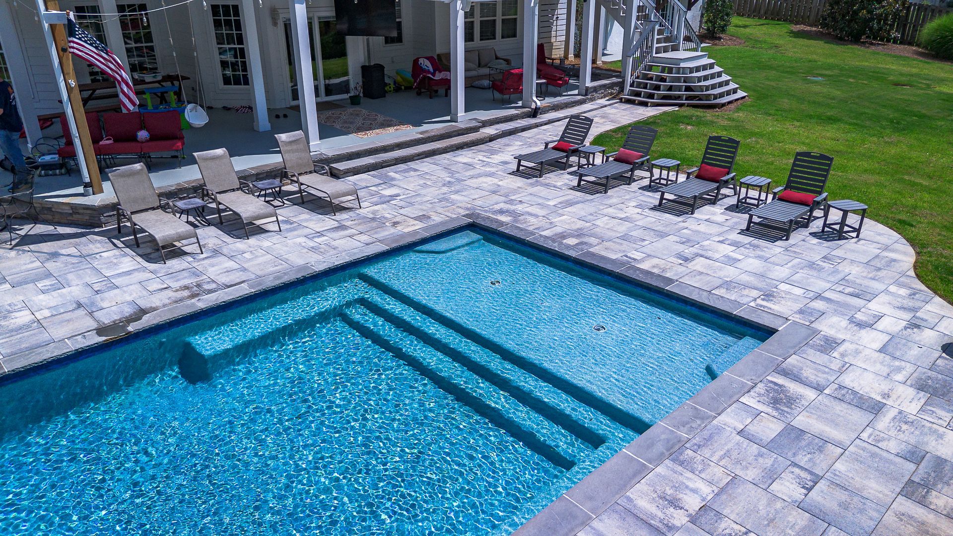Pool with blue water and steps, surrounded by a patio, lounge chairs, and a house.