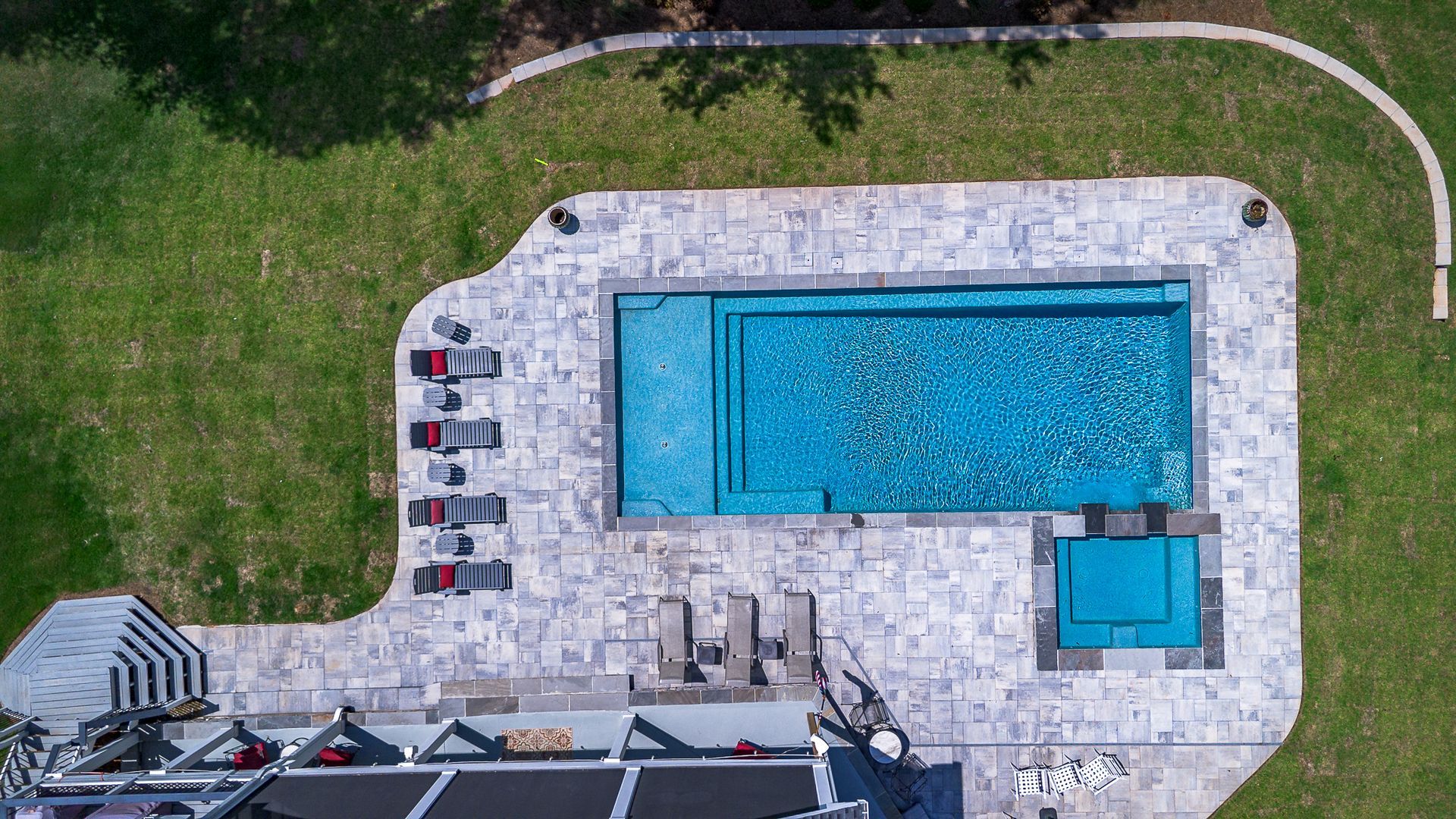 Overhead view of a rectangular pool, surrounded by stone patio and grassy yard. Lounge chairs, small hot tub.