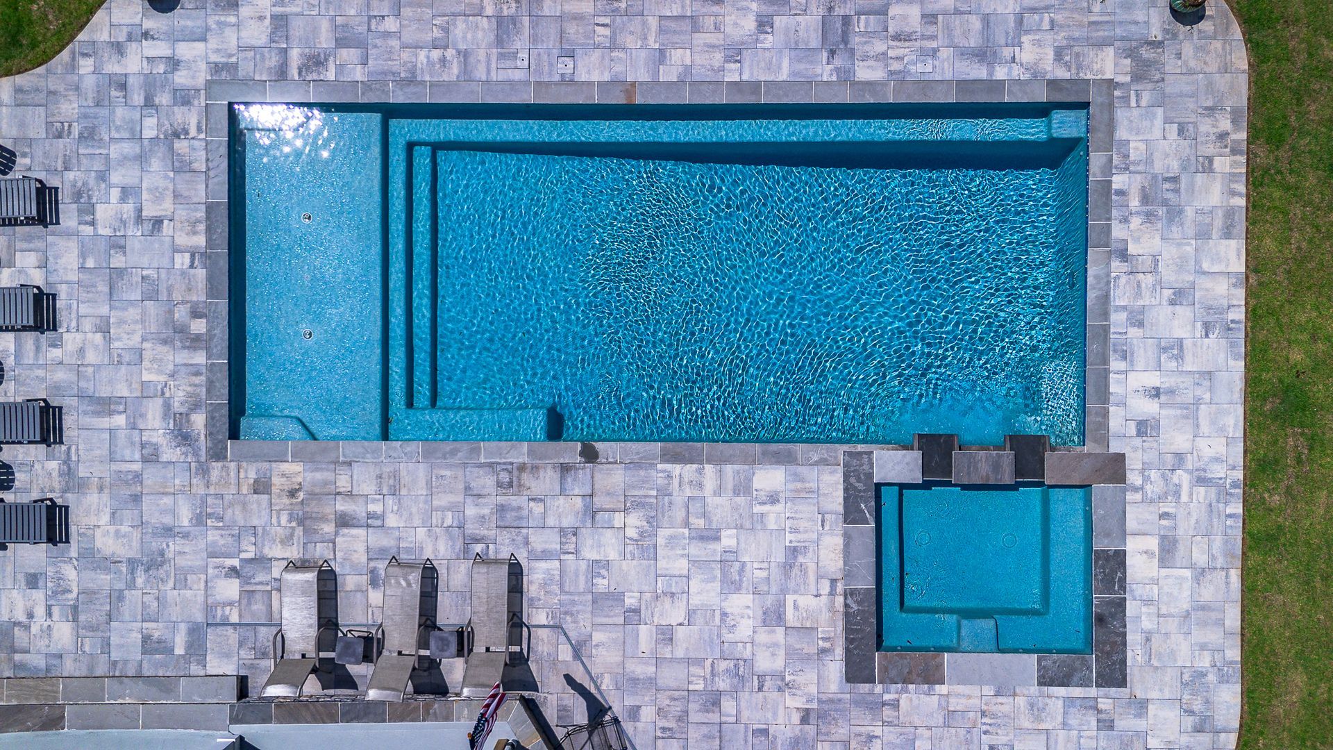 Aerial view: rectangular pool with a spa, surrounded by gray stone patio. Bright blue water.