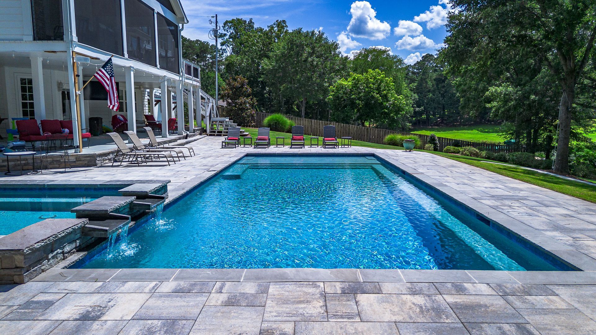 A large rectangular swimming pool in a backyard with a white house, blue sky and green trees.