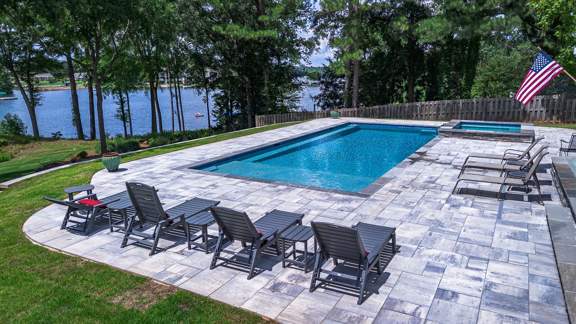 Poolside scene overlooking a lake. Gray patio with lounge chairs, a pool, and a jacuzzi. American flag.