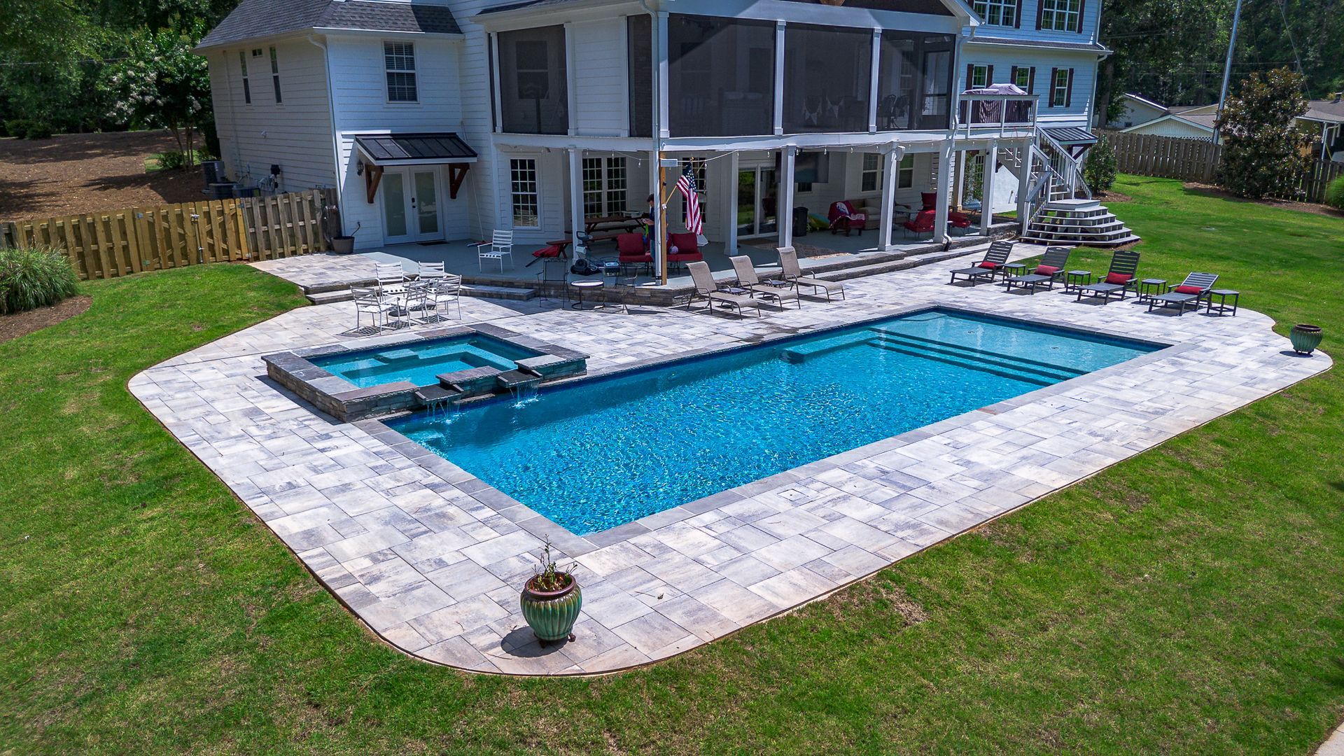 Backyard with a rectangular pool and hot tub surrounded by stone patio; two-story white house in the background.