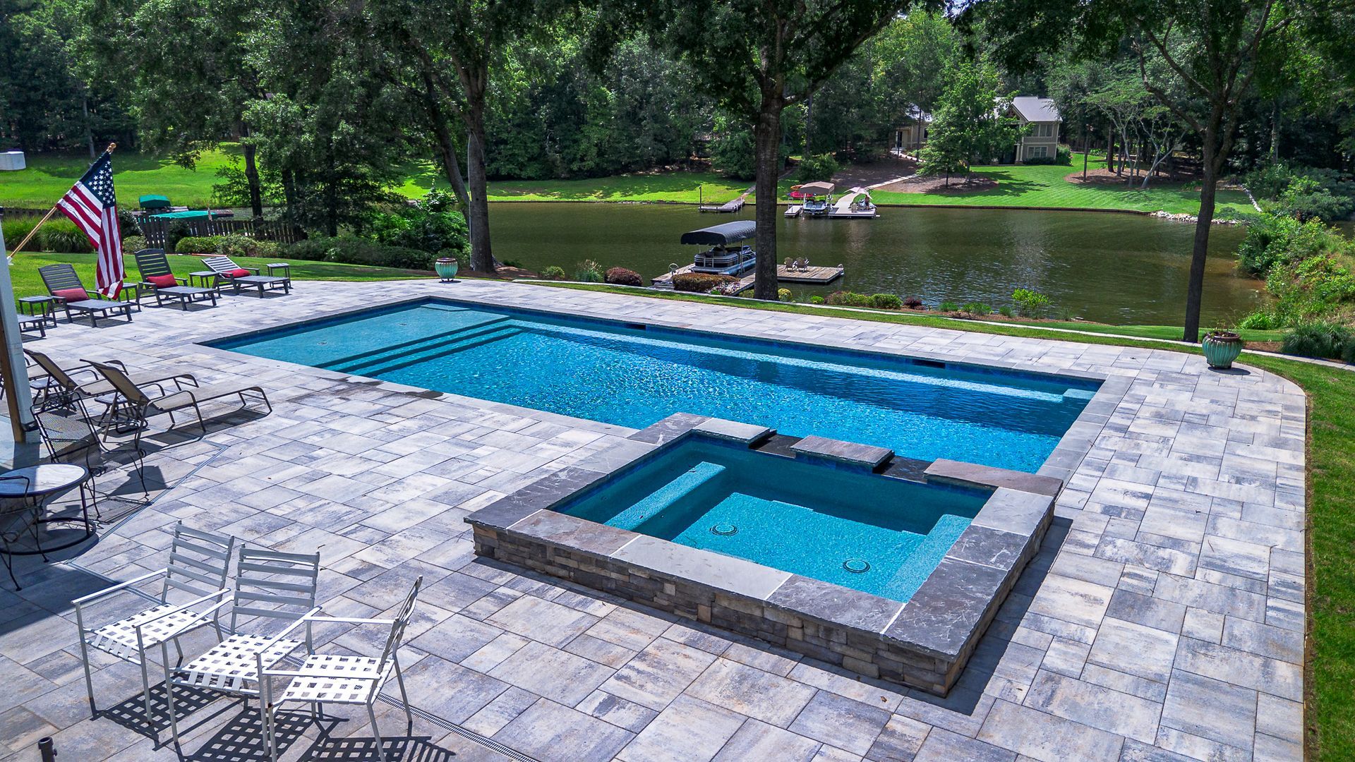 Pool and spa on a patio overlooking a lake, with trees and an American flag.