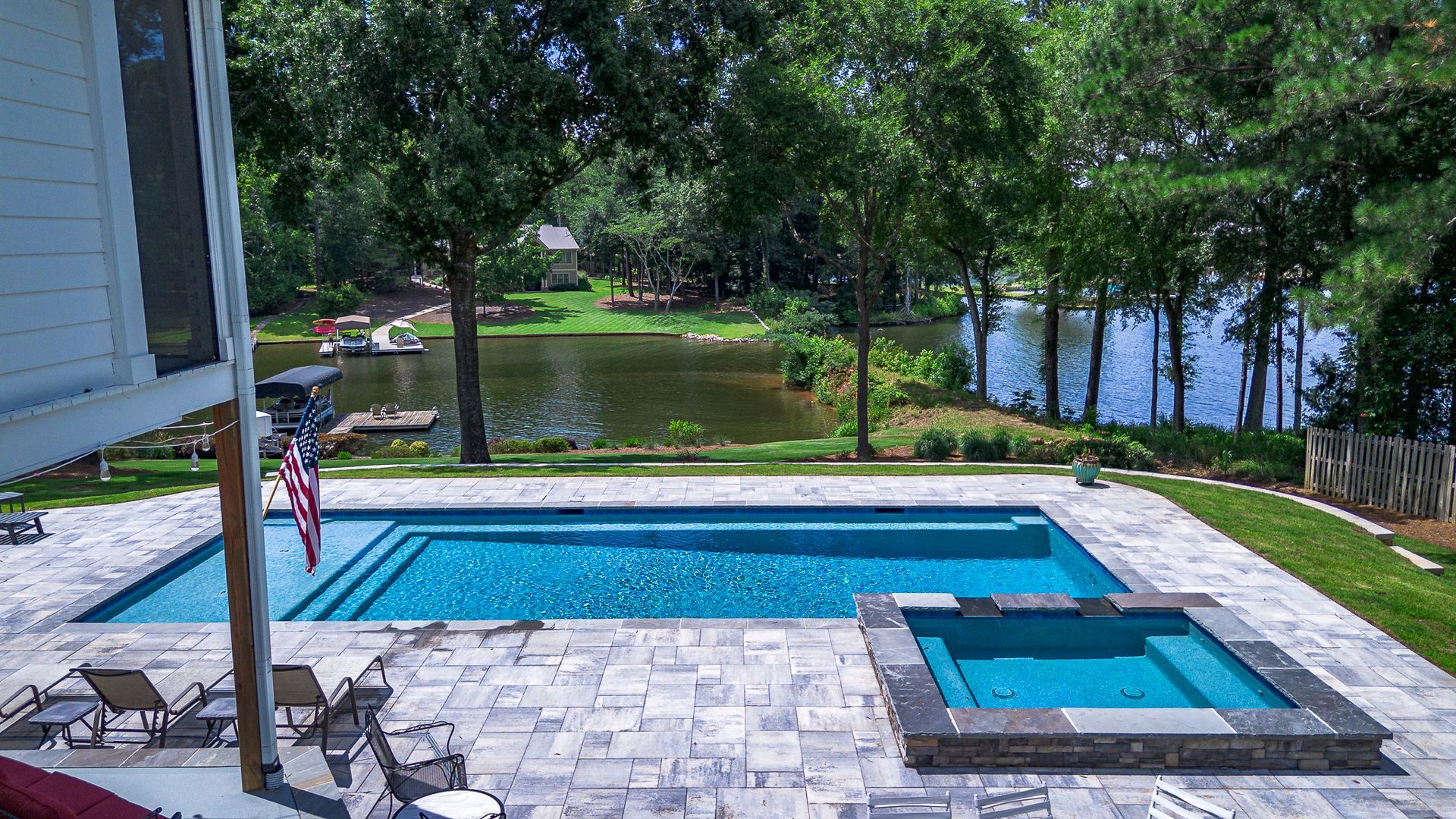 Pool with lake view: blue water, flag, chairs, trees, sunny day.