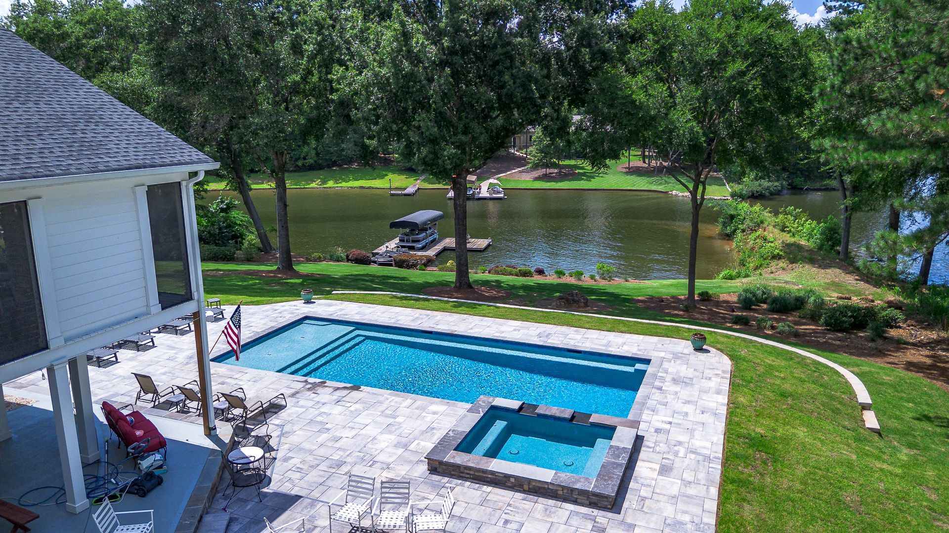 Poolside view: blue pool and spa next to a lake with trees, house on the left.