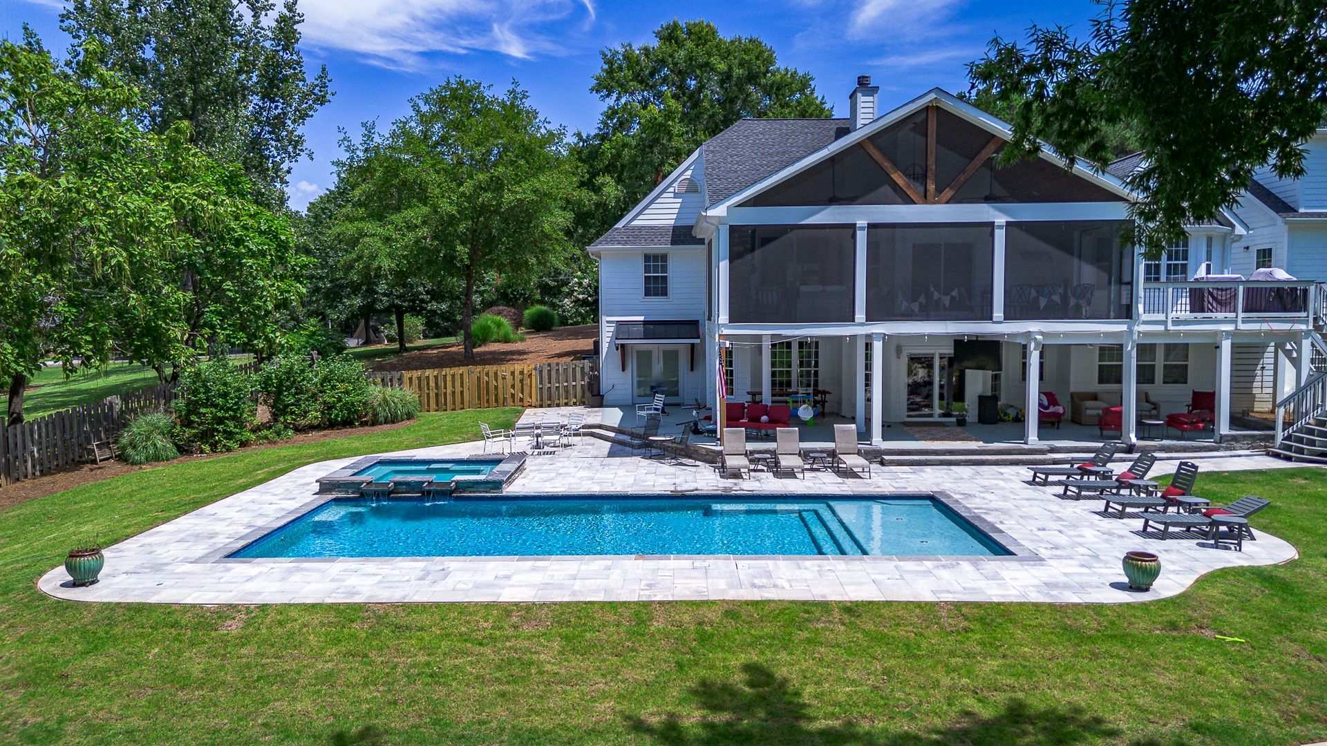 Backyard of a white house with pool, screen porch, lounge chairs, and lush green lawn. Sunny day.