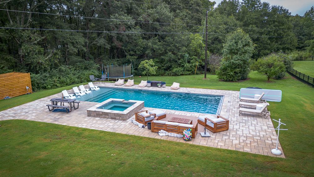 Backyard pool with hot tub, lounge chairs, and seating area surrounded by green grass and trees.