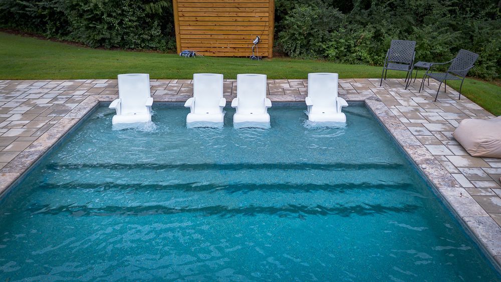 Swimming pool with four white in-pool chairs, steps, and patio seating, set against green lawn and foliage.