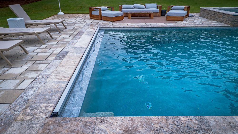 Pool with patio furniture. Light blue water, stone patio, wooden furniture, and green grass.
