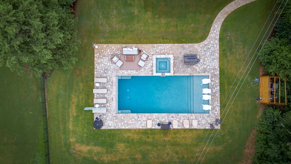 Overhead view of a rectangular pool with a hot tub, patio, lounge chairs, and green lawn.