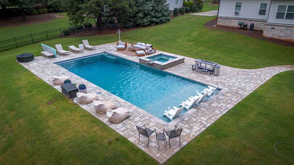 A rectangular pool surrounded by stone patio, lounge chairs, and hot tub in a grassy backyard.