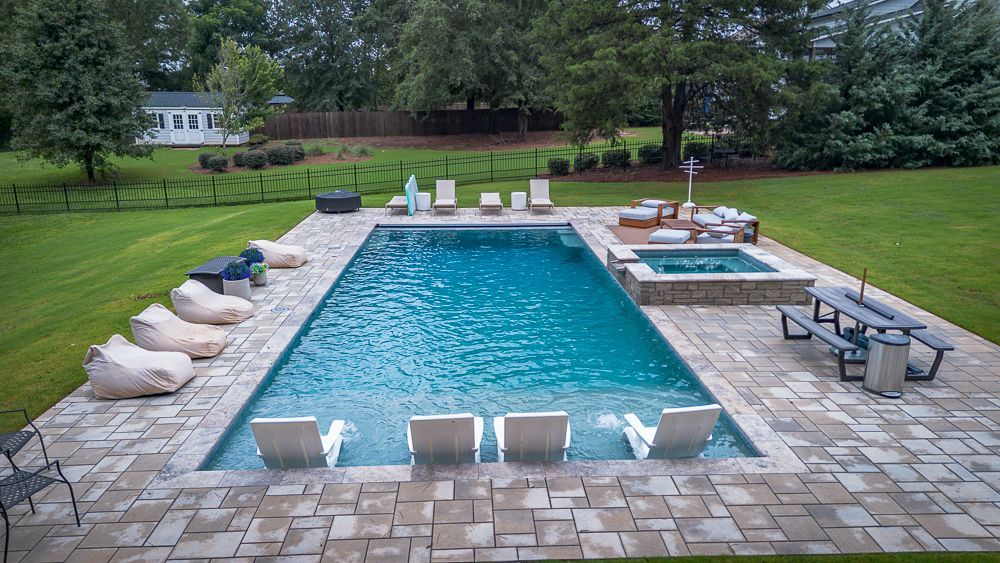 Backyard pool with a hot tub, lounge chairs, and bean bag chairs on a patio. Green grass surrounds the pool.