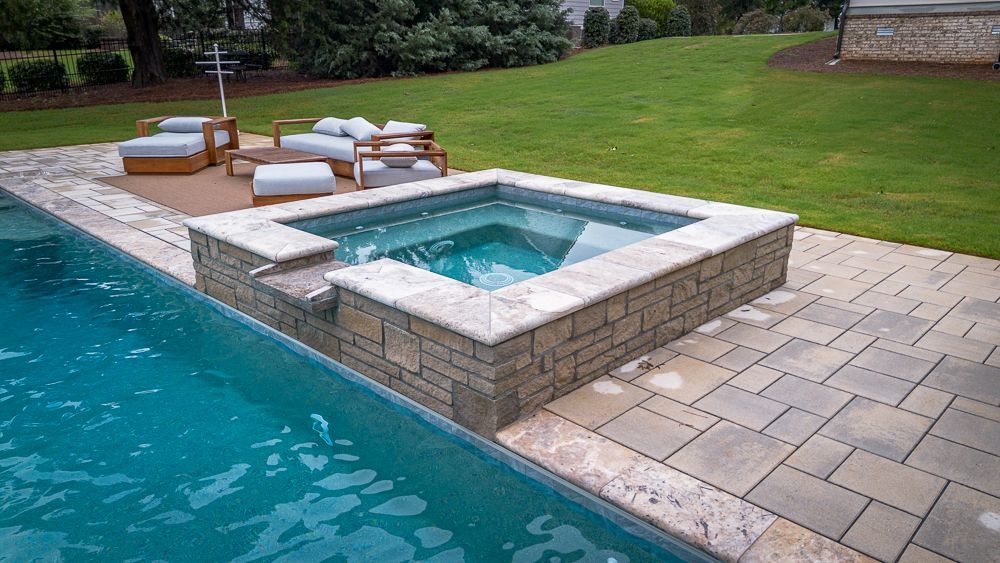 Hot tub and pool surrounded by stone patio in a backyard with seating area and green grass.