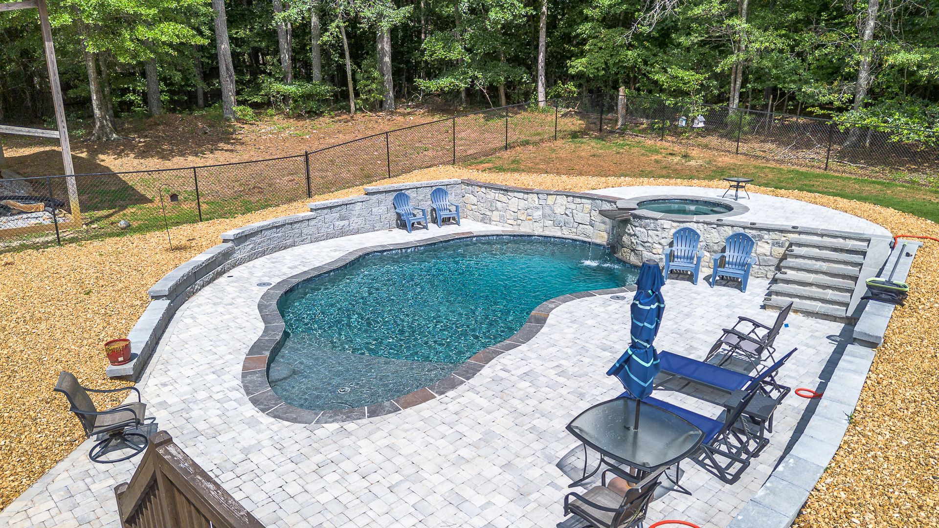 A backyard pool with stone patio, hot tub, seating, and water features. Trees in the background.