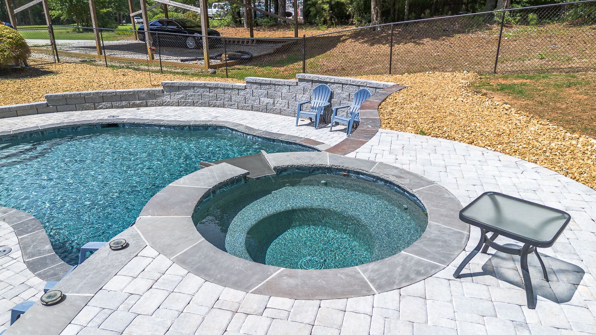 Swimming pool with integrated hot tub, patio, and chairs. Gray stone, blue water.