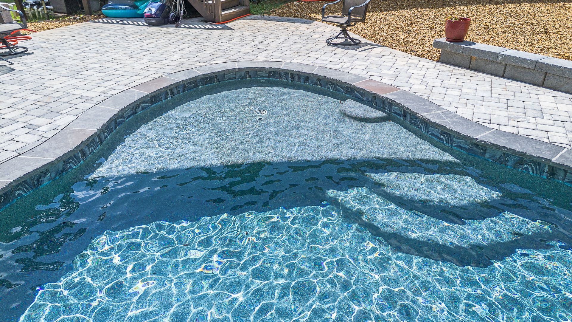 Swimming pool with stone coping and steps, blue water reflecting sunlight, surrounded by pavers.
