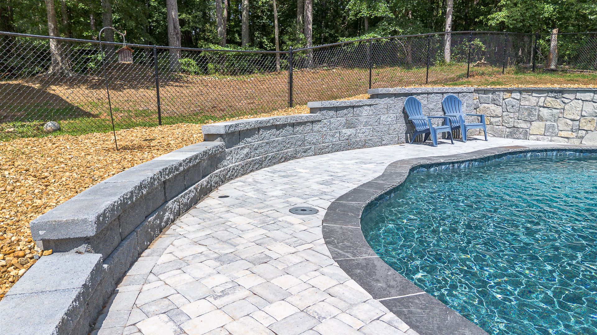 Curving pool with stone pavers, retaining wall, and two blue chairs. Wooded area in the background.
