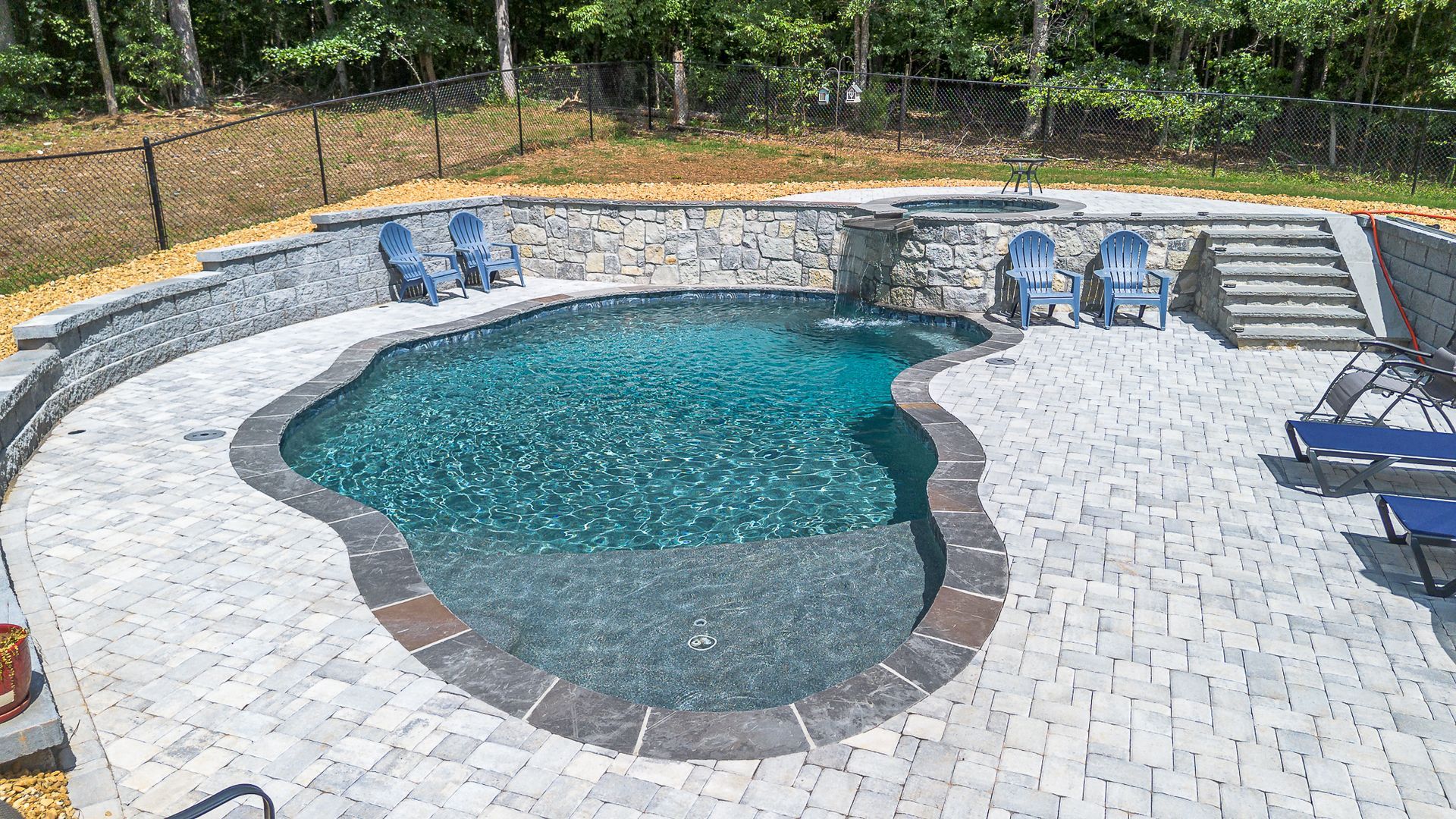 Pool with waterfall feature, stone patio, blue chairs, and steps.