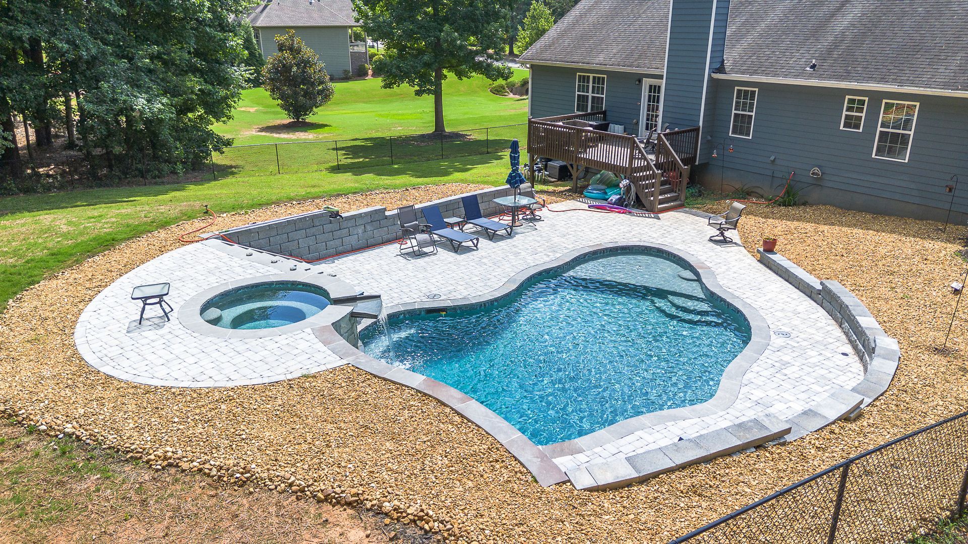 Backyard pool with spa, blue water, white paving, brown gravel border, house in background.