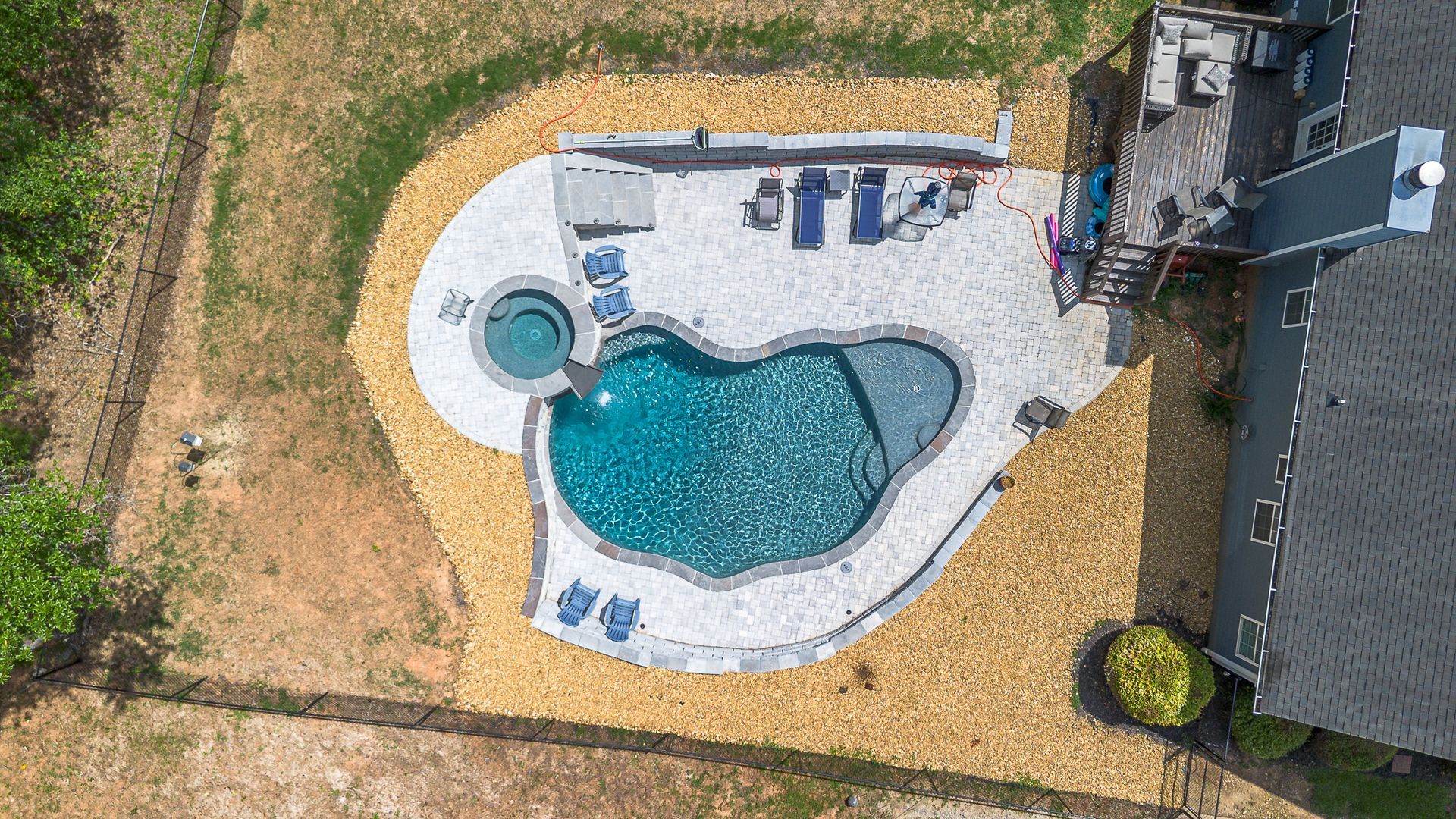 Aerial view of a backyard pool with a hot tub, patio, and deck, surrounded by landscaping and a grassy yard.