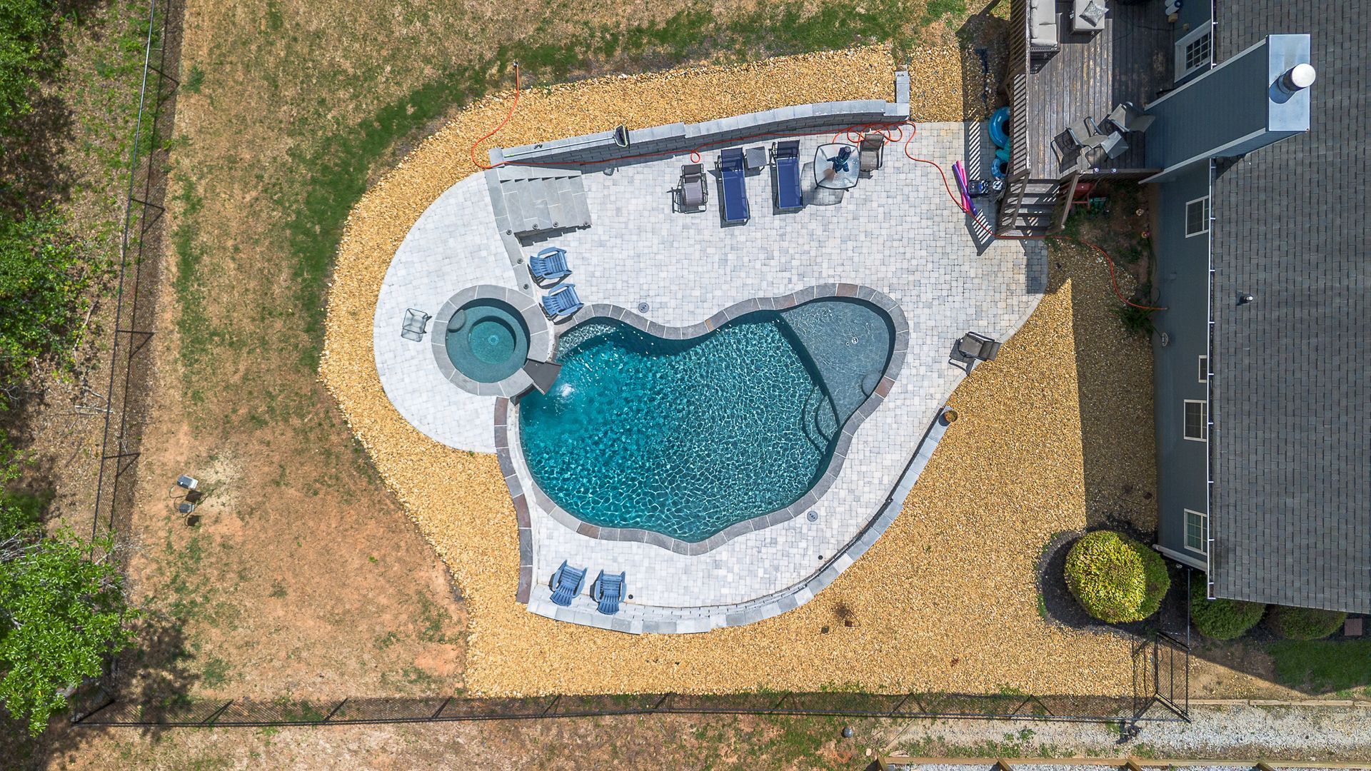 Aerial view of a pool and hot tub surrounded by stone, near a house and grassy yard.