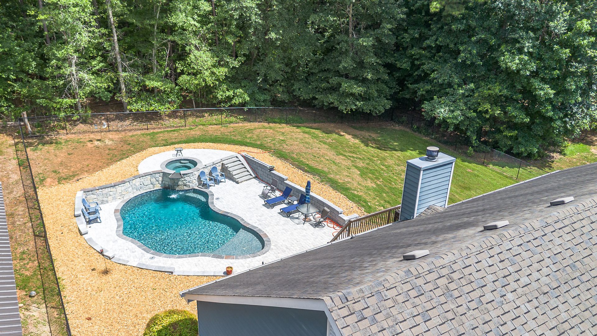 Aerial view of a backyard pool with a hot tub, surrounded by patio and lawn, near a house and forest.