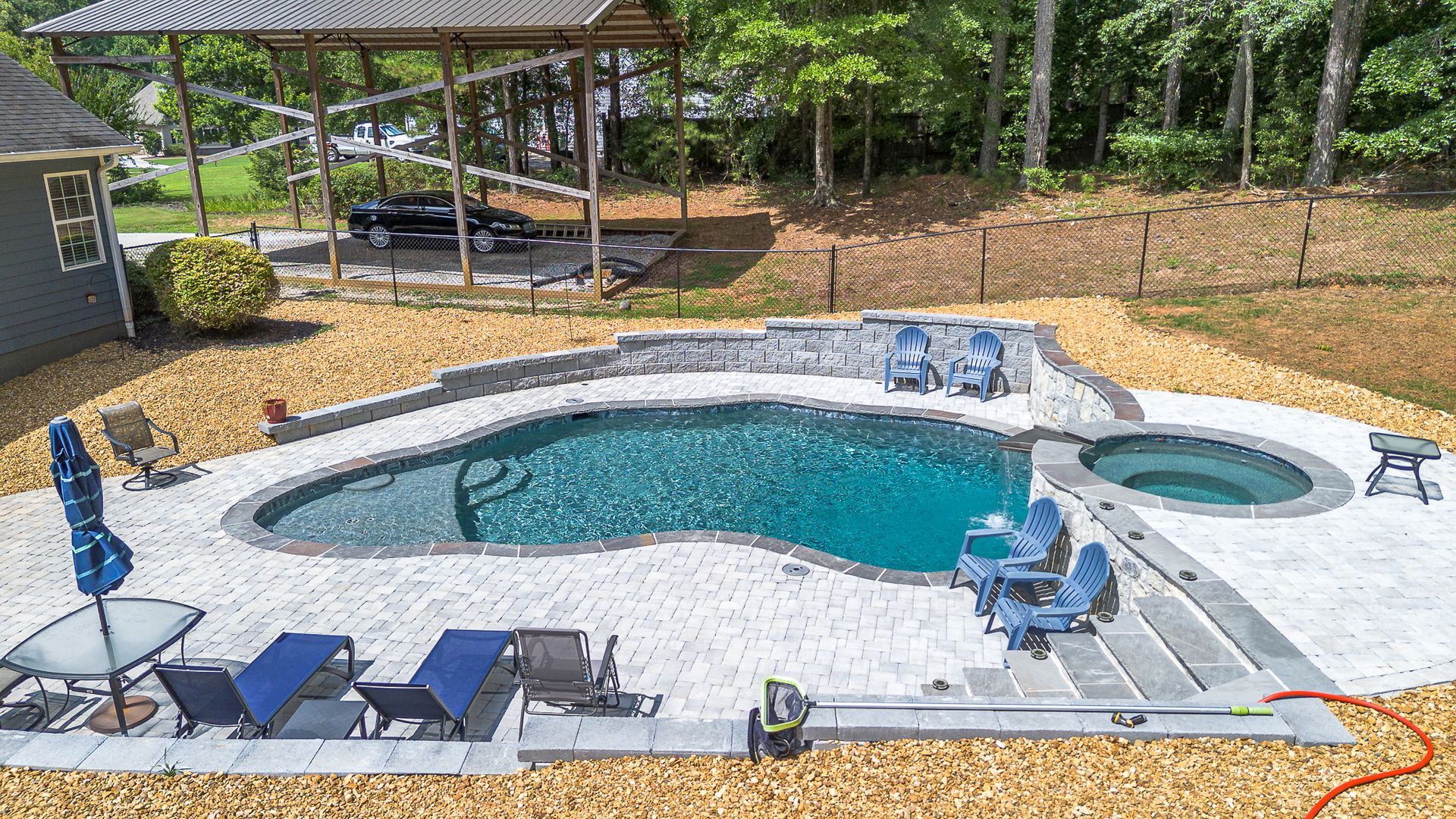 Backyard pool with a hot tub, chairs, and a grill, set on gray stone and surrounded by brown mulch.
