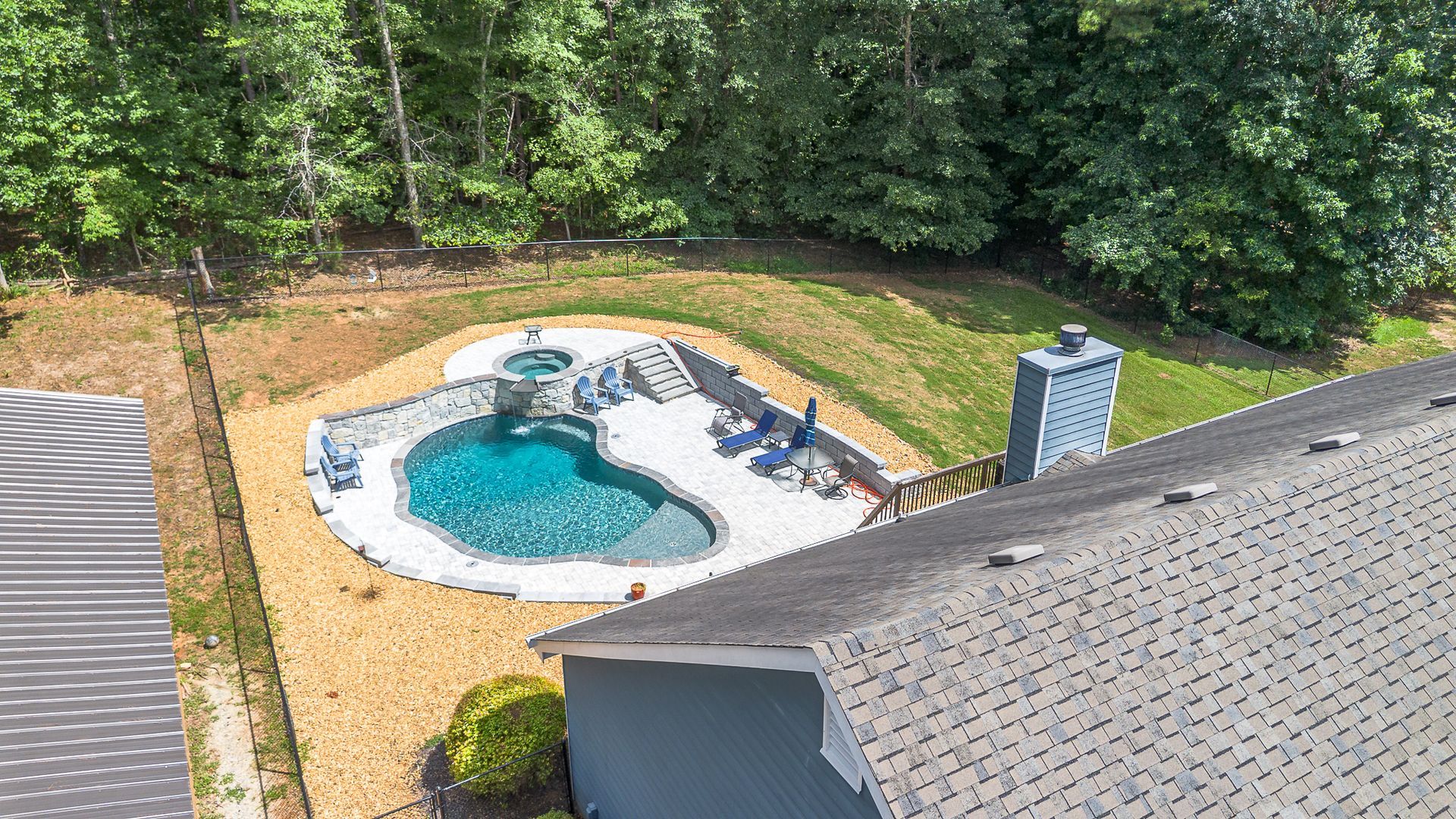 Aerial view of a pool with a waterfall, surrounded by a rock patio and lawn, next to a house with a gray roof.