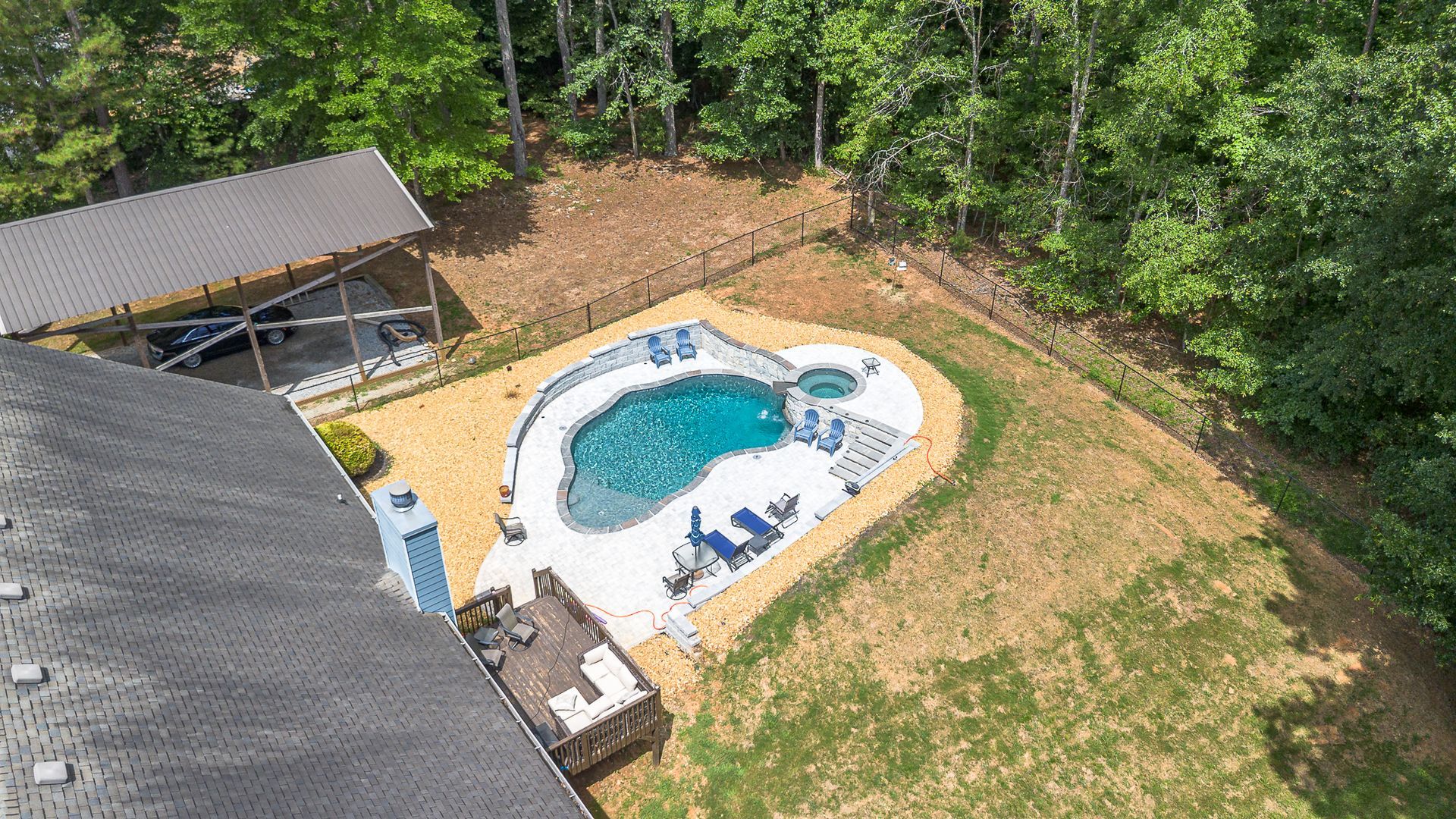 Aerial view: Heart-shaped pool surrounded by lawn and trees; a carport, house, and small deck are also present.