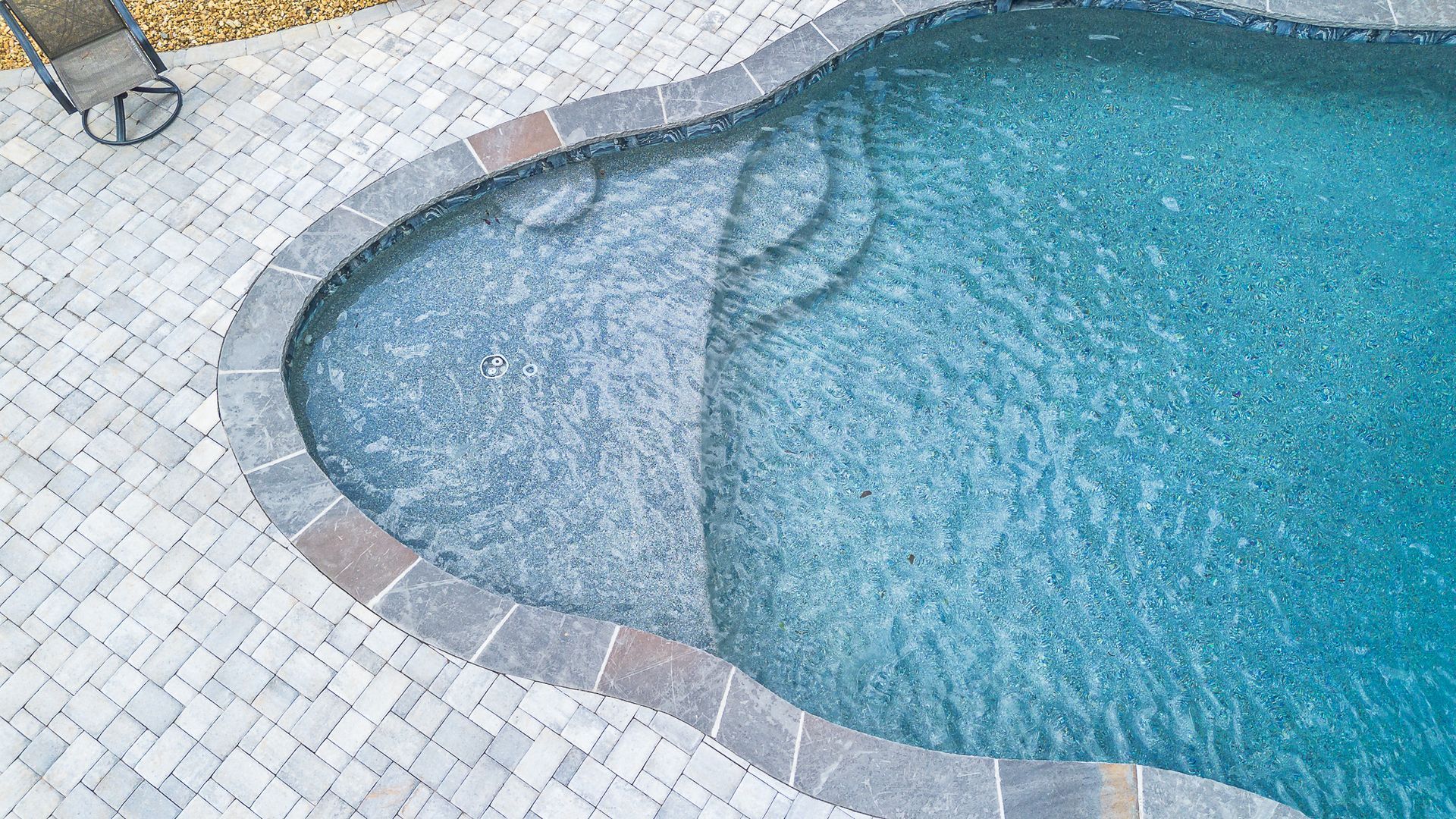 Aerial view of a swimming pool with steps, surrounded by gray pavers and a small chair.